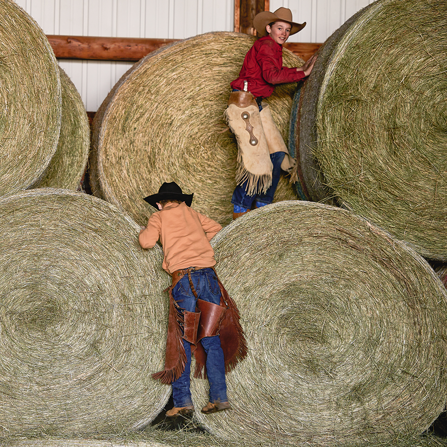 Two boys dressed as cowboys climbing on big round bales of hay running from each other.